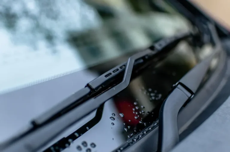 Windshield wiper blade resting at the base of a car windshield with water drops on the cowl