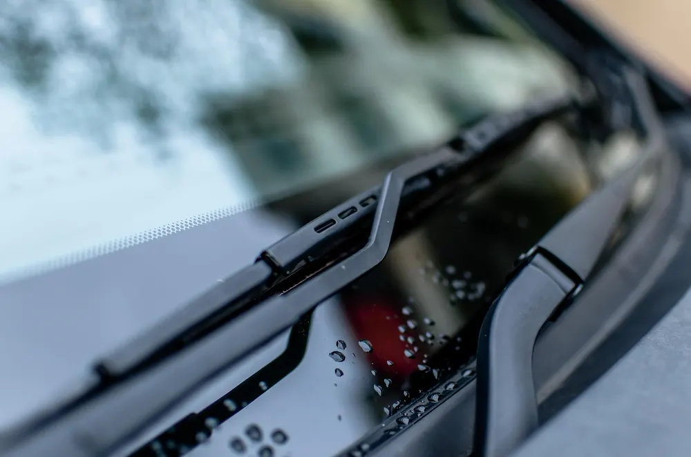 Windshield wiper blade resting at the base of a car windshield with water drops on the cowl