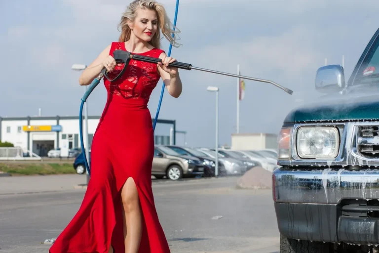 Person in a red dress aiming a pressure washer lance at the front of a truck in a parking area