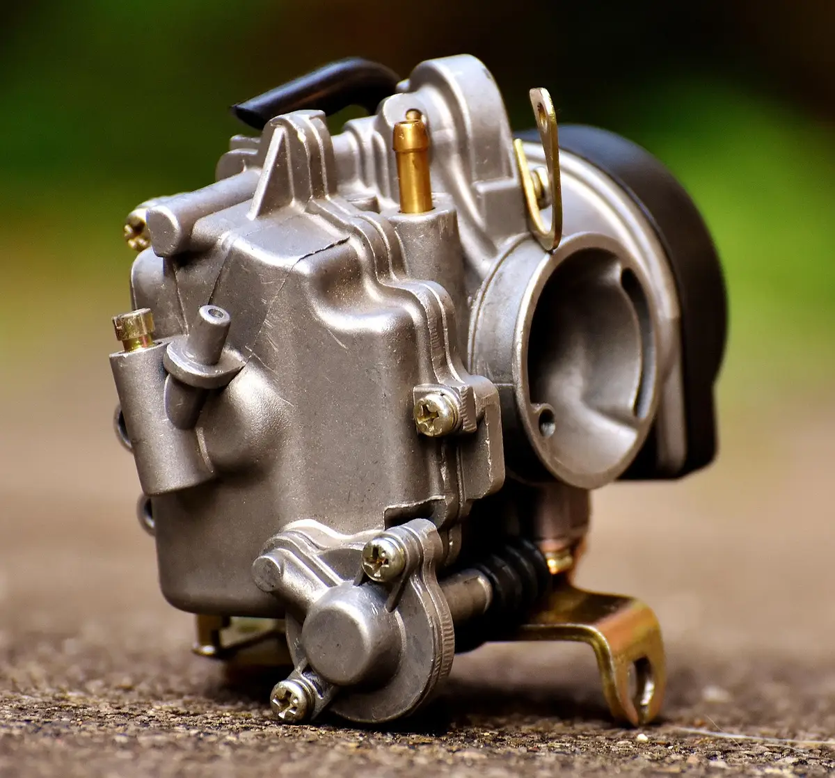 Small engine carburetor body with throttle bore, fuel bowl and linkage on a gravel surface
