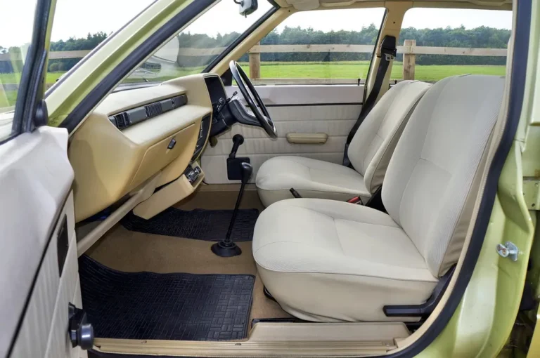 Open door of a vintage car showing beige door panel, seats and rubber floor mats