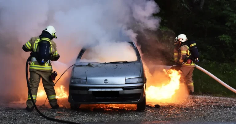 Firefighters in protective gear spraying water on a burning passenger car beside a road