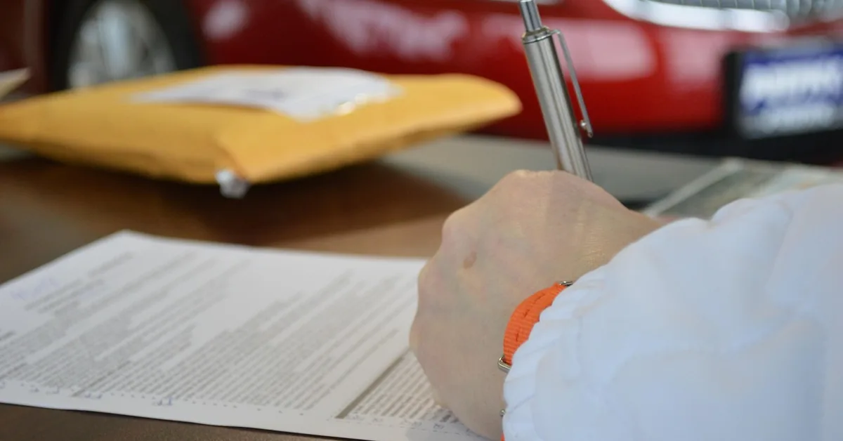 Hand signing vehicle sale paperwork at a desk with a red car behind
