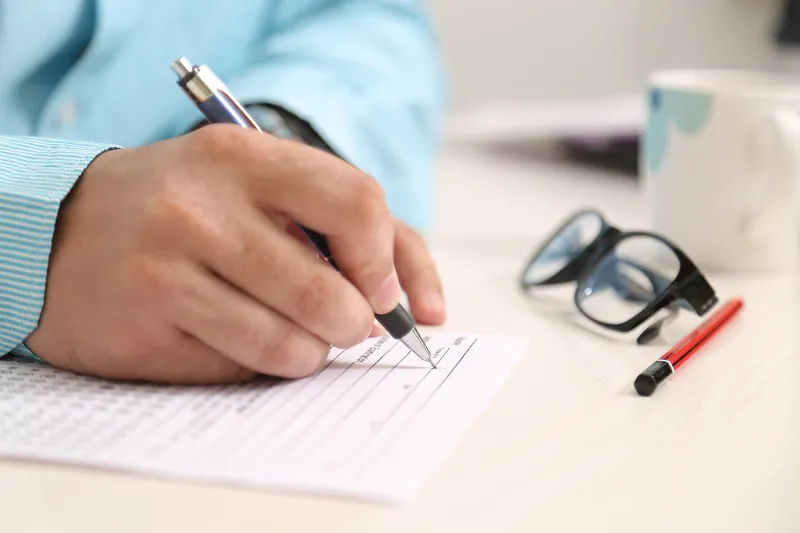Hand holding a pen over a paper form with rows of boxes on a desk, next to a pair of glasses