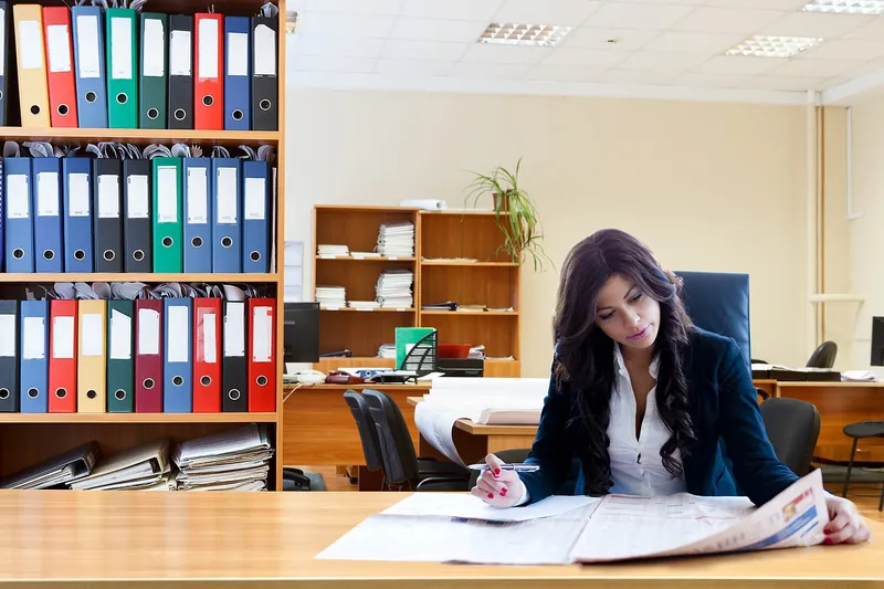 Woman at an office desk filling in a paper form beside shelves of document binders