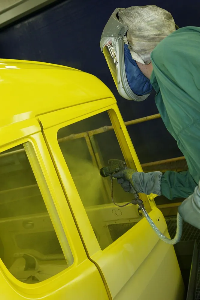 Painter in a hooded face shield spraying a yellow vehicle panel