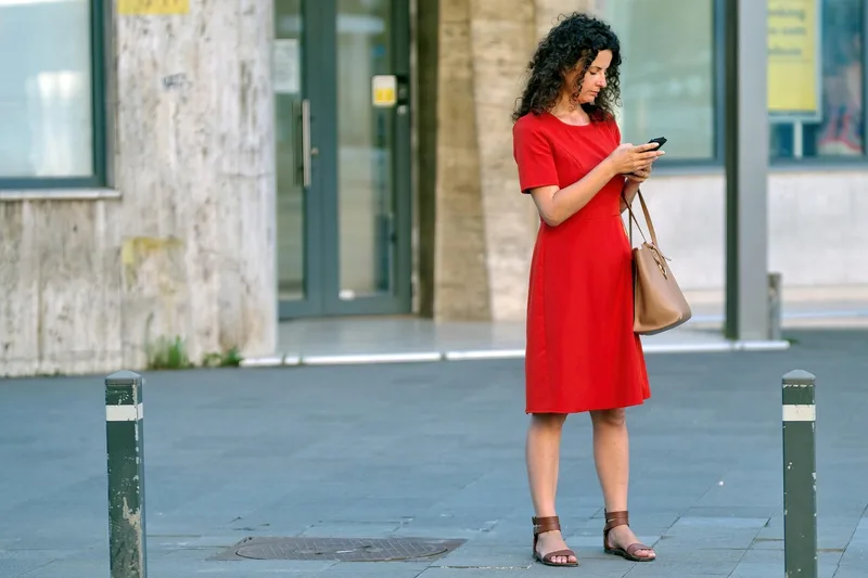 Woman in a red dress checking a smartphone while standing on a city sidewalk