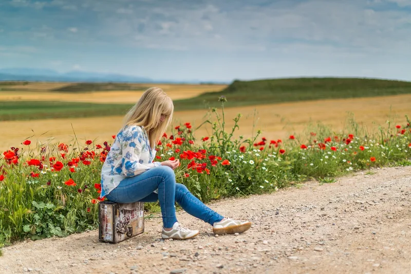 Woman sitting on a small suitcase beside a country road with poppies