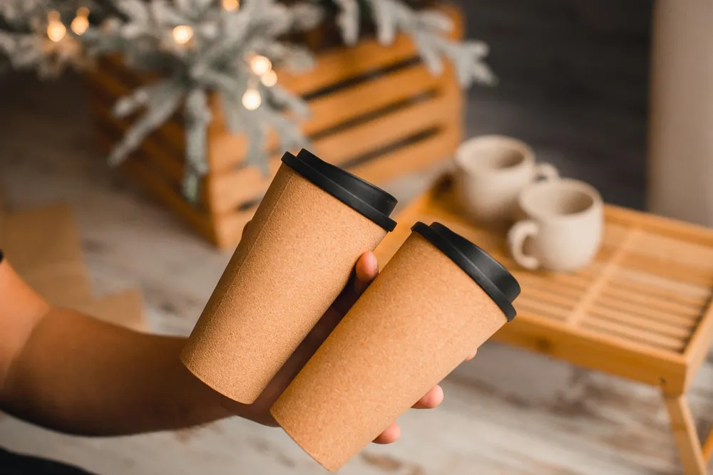 A hand holding two cork-textured travel cups with black lids