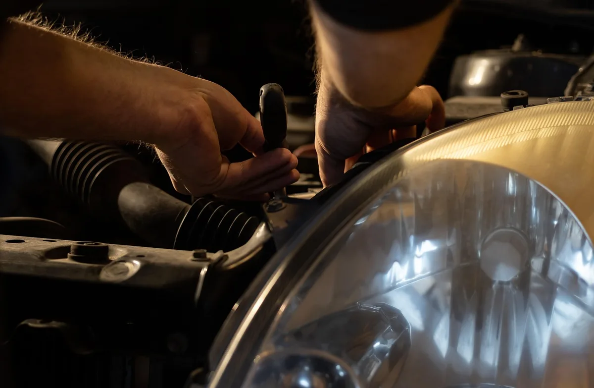 Mechanic’s hands working on a car engine bay near a headlight assembly, checking a wiring connector