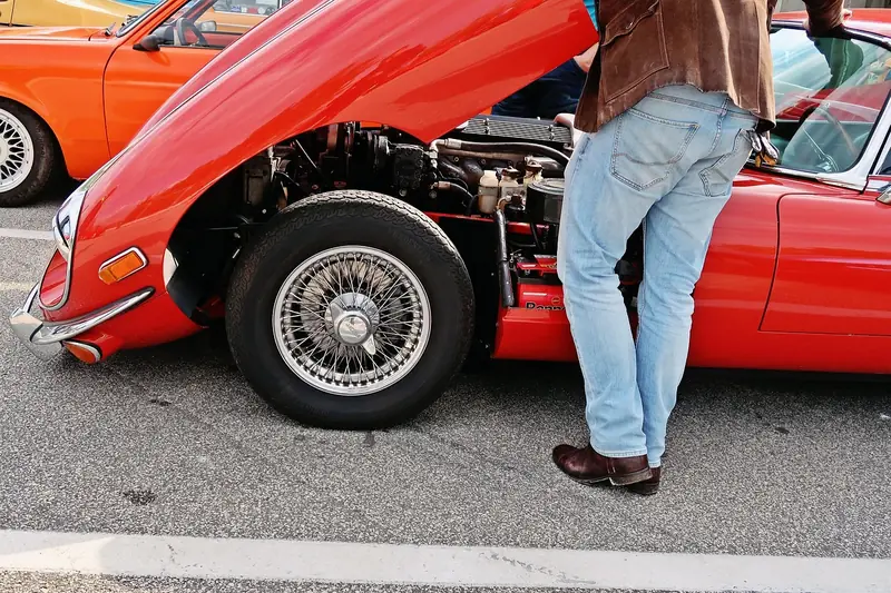 Person standing at the open hood of a red classic car in a parking area
