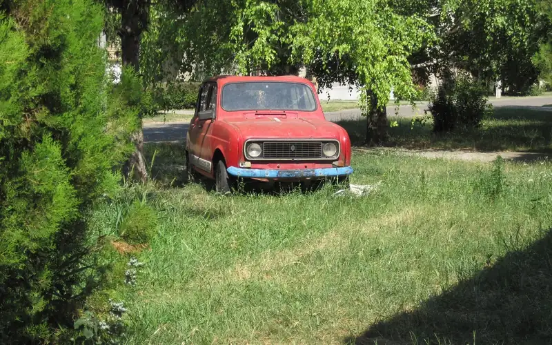 Older red hatchback parked on grass under trees Older red hatchback parked on grass under trees
