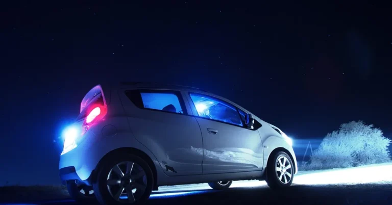Silver hatchback with its headlights and tail light on at night under a starry sky