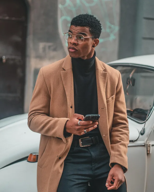 Man in a camel coat checking his phone while leaning on a parked white car