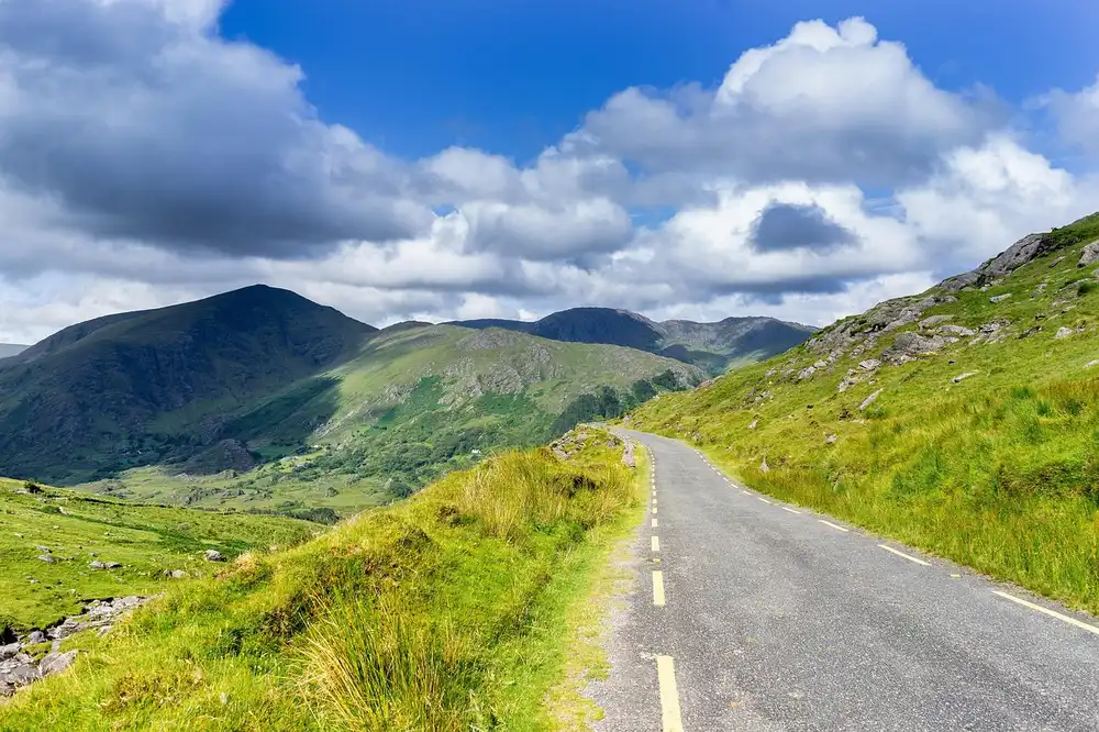 A narrow paved road with a yellow edge line running along a green hillside under a cloudy blue sky