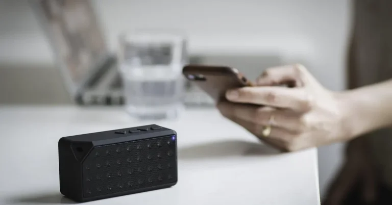 Smartphone held beside a small black Bluetooth speaker on a white table