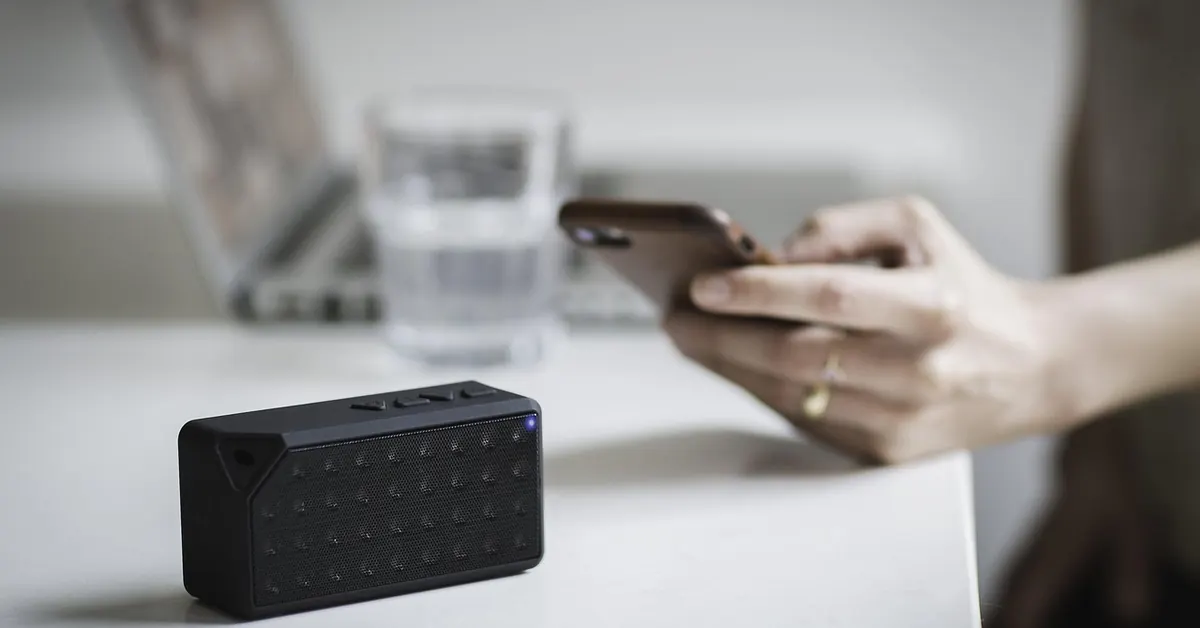 Smartphone held beside a small black Bluetooth speaker on a white table