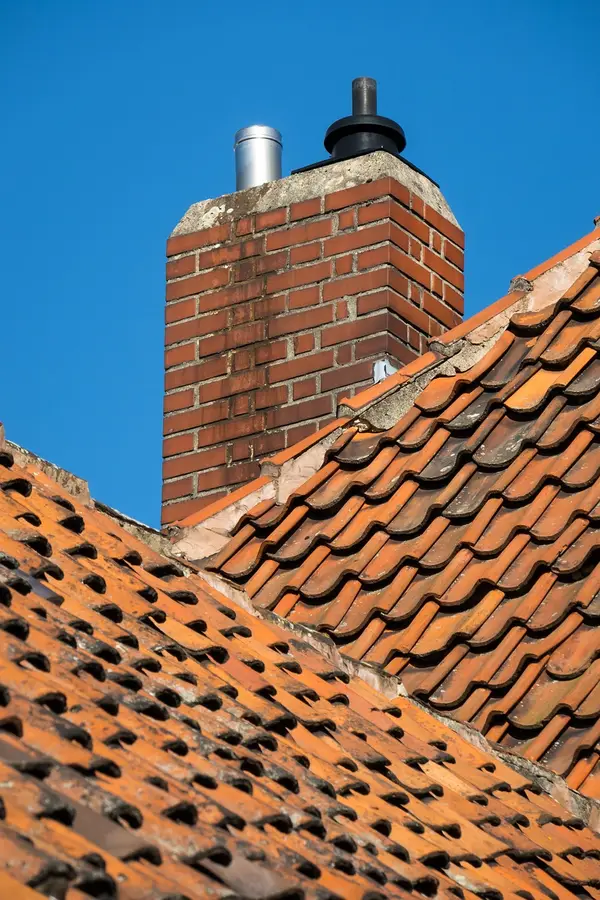 Brick chimney with two vent pipes on a tiled roof against a blue sky