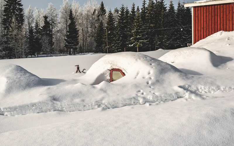 Car buried under deep snow next to a red barn