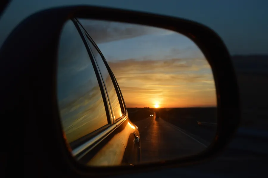 Side mirror of a car showing an open road and the setting sun behind it