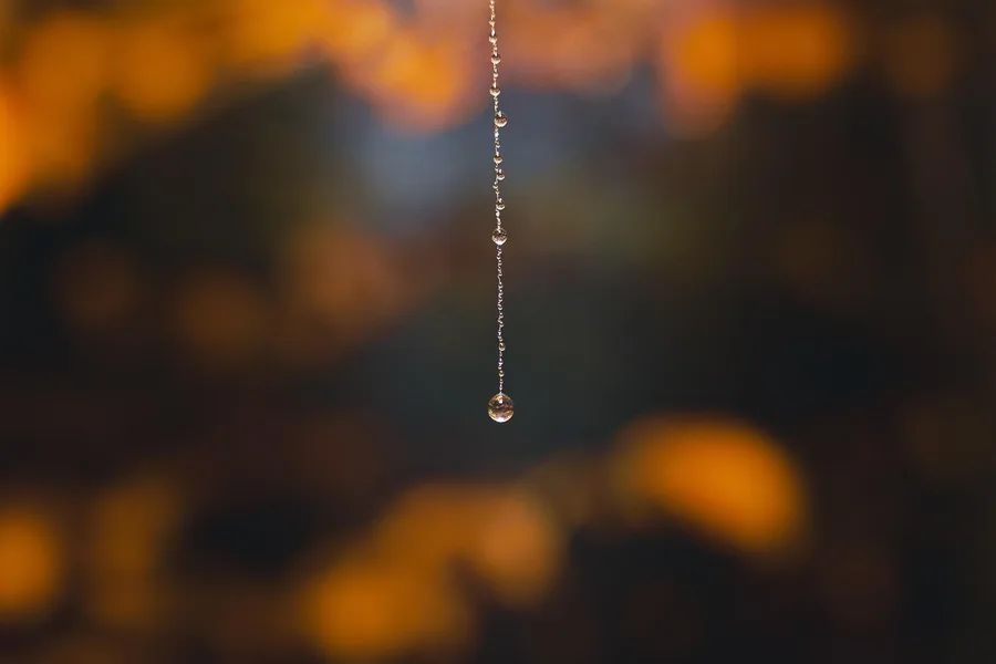A single water drop hanging from a thin thread against a blurred background
