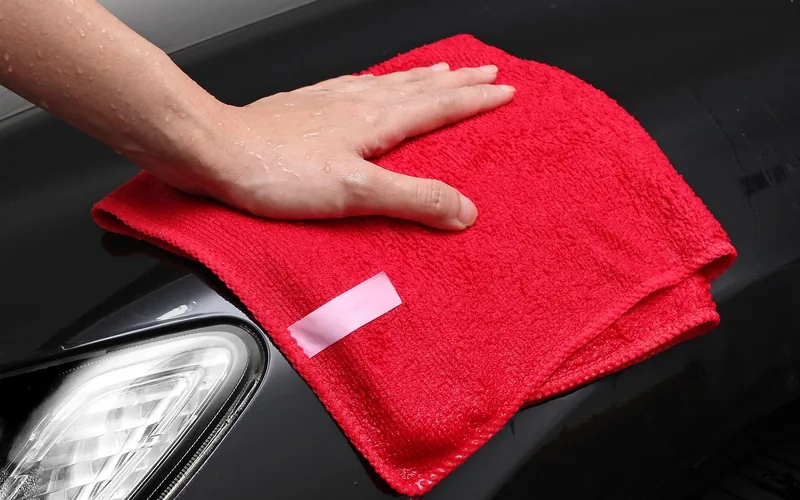 A hand wiping a black car body panel next to the headlight with a red microfiber towel
