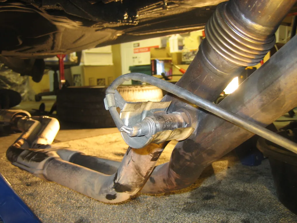 Gloved hands installing an oxygen sensor into an exhaust pipe under a car