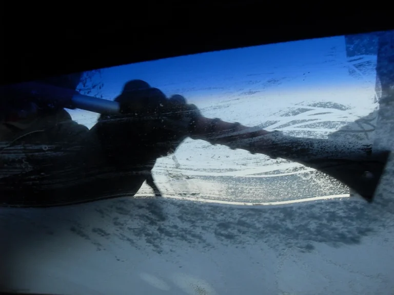 Driver with de-icer spray and scraper beside a frost-covered windshield at sunrise