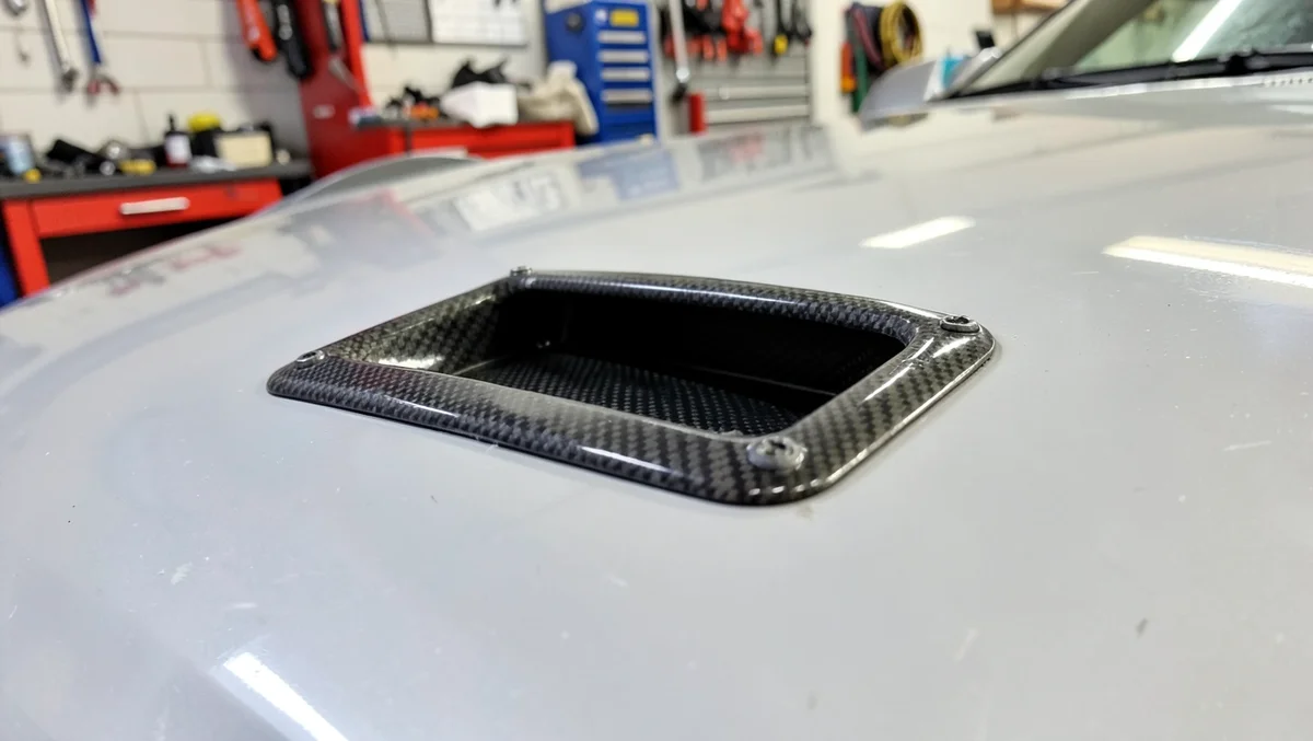 Carbon fiber decorative hood scoop bolted onto a car hood in a home garage