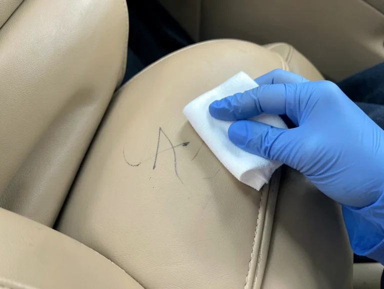 Gloved hand dabbing an ink stain on a tan leather car seat with a cotton pad