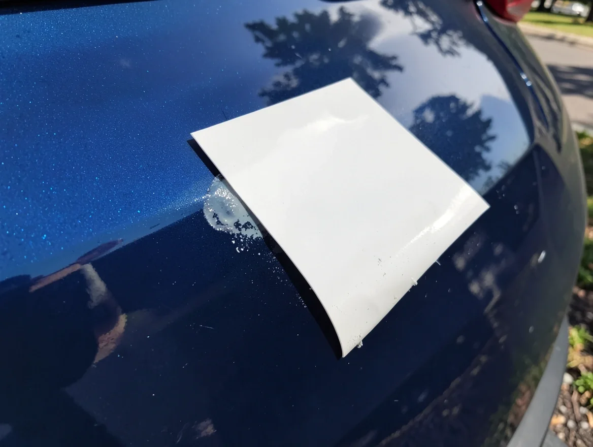 Plastic scraper card lifting the corner of a dealership decal off glossy car paint, showing leftover adhesive residue