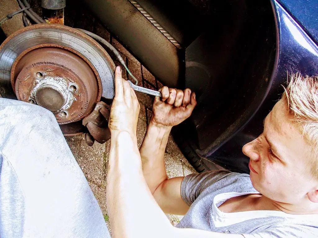Mechanic and car owner inspect a sedan with open hood, battery, coolant reservoir, and tire in a garage