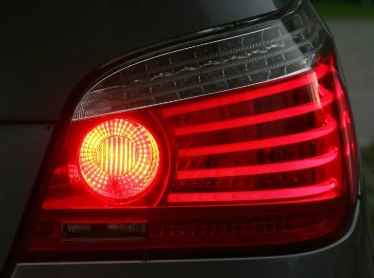 Mechanic compares car brake light bulbs beside a sedan in a daylight garage.
