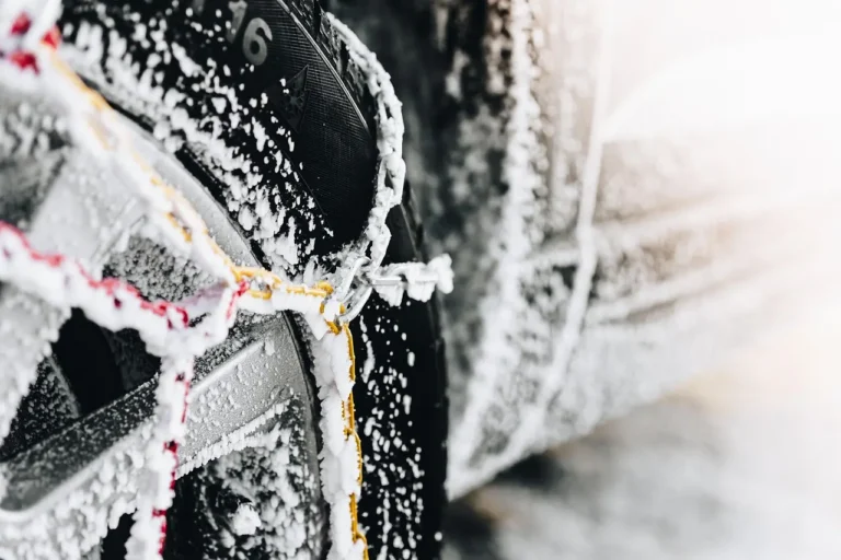 Modern sedan in snow while a driver checks tire chain fitment near the front wheel