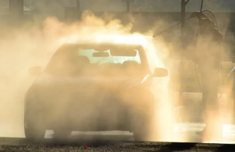 Person washing a dark sedan with foam cannon and buckets in natural light