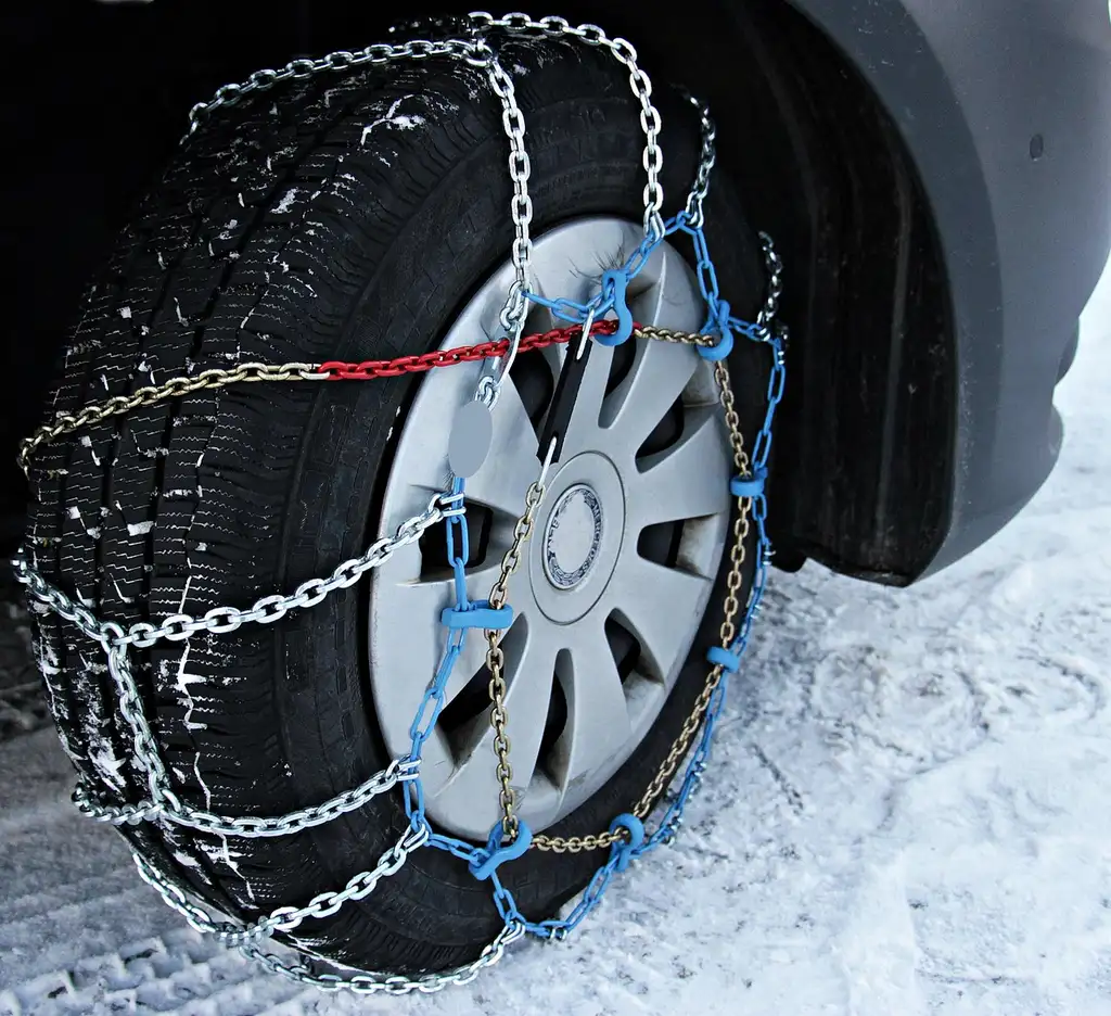 Driver fitting car snow chains beside a compact SUV on a snowy mountain roadside
