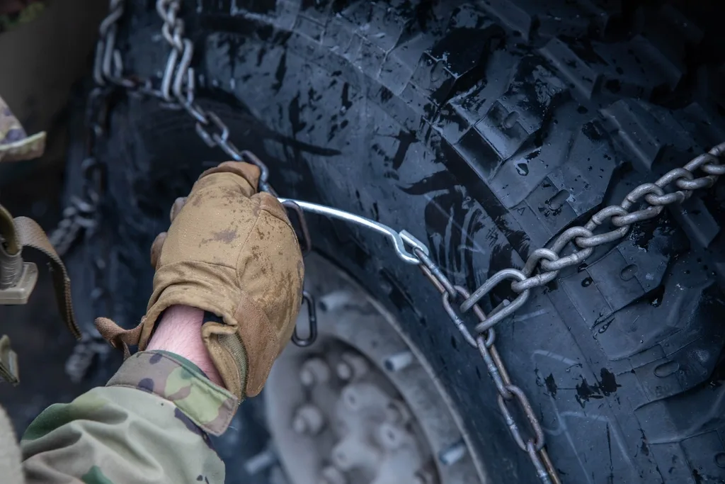 Gloved hands tightening a car snow chain tensioner on a tire in snow