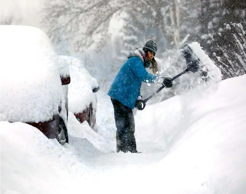 Gloved hands use a metal snow shovel to break up ice packed under a car tire.