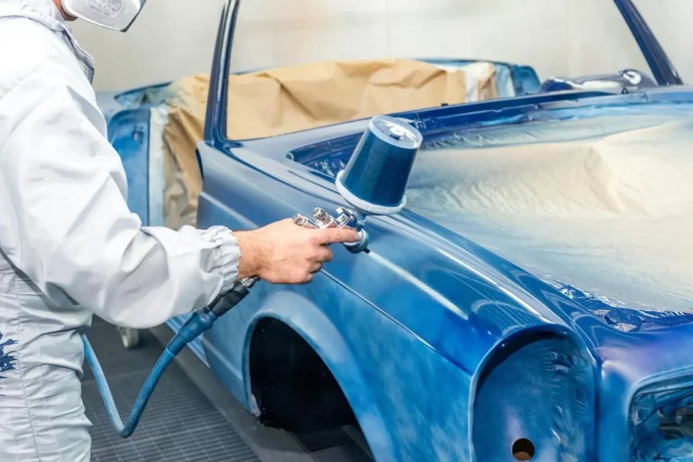 Mechanic comparing painted plastic trim samples beside a car in a sunlit garage