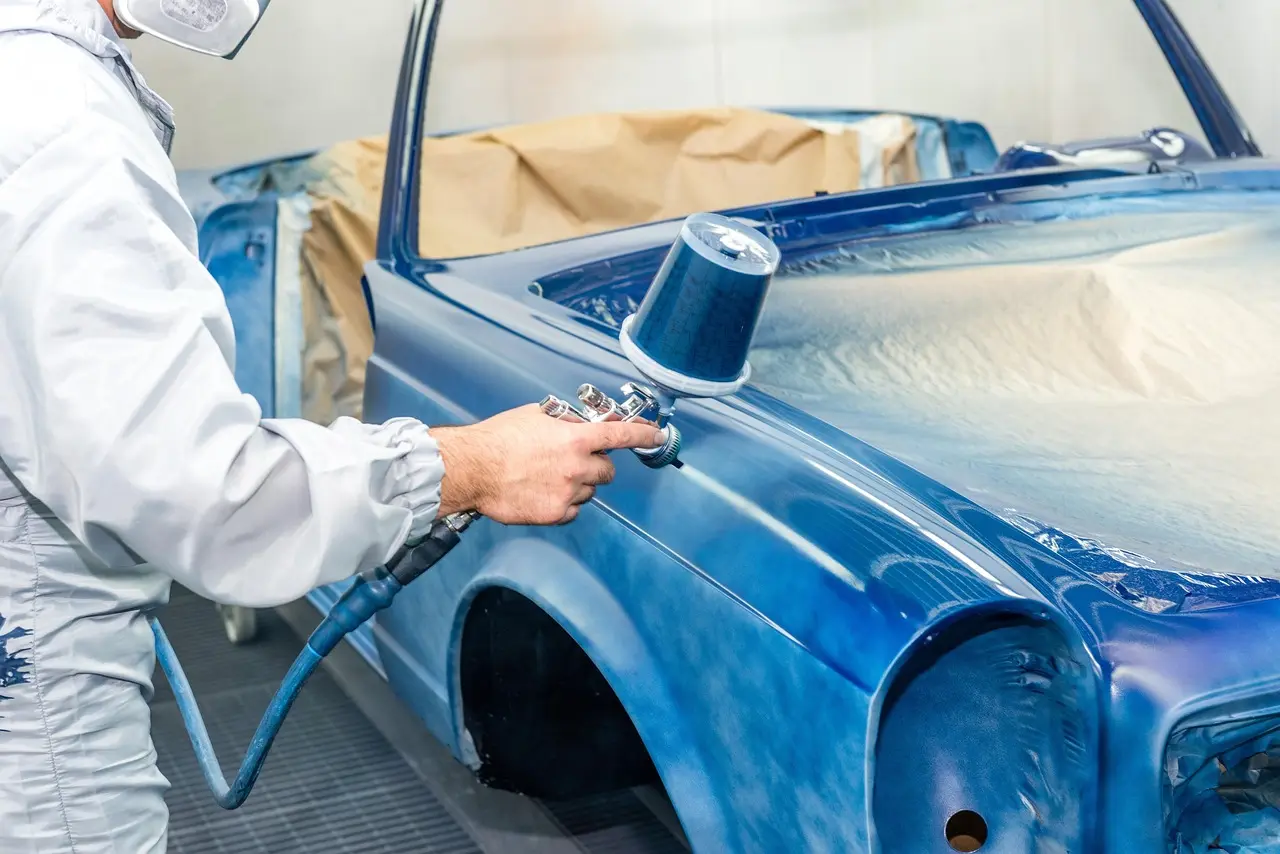 Mechanic comparing painted plastic trim samples beside a car in a sunlit garage