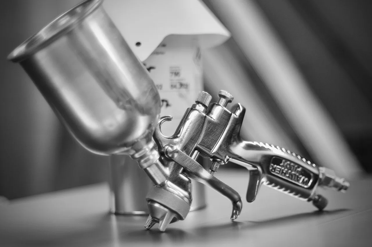 Three car paint sprayers on a workbench beside a fender in a naturally lit garage