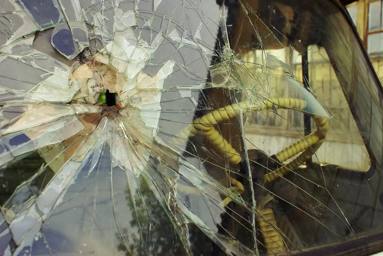 A car owner examines a cracked windshield beside a parked sedan in daylight.