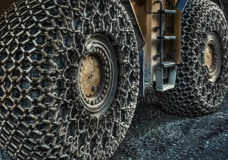 Driver fits snow chains onto a car tire on a snowy roadside in daylight.