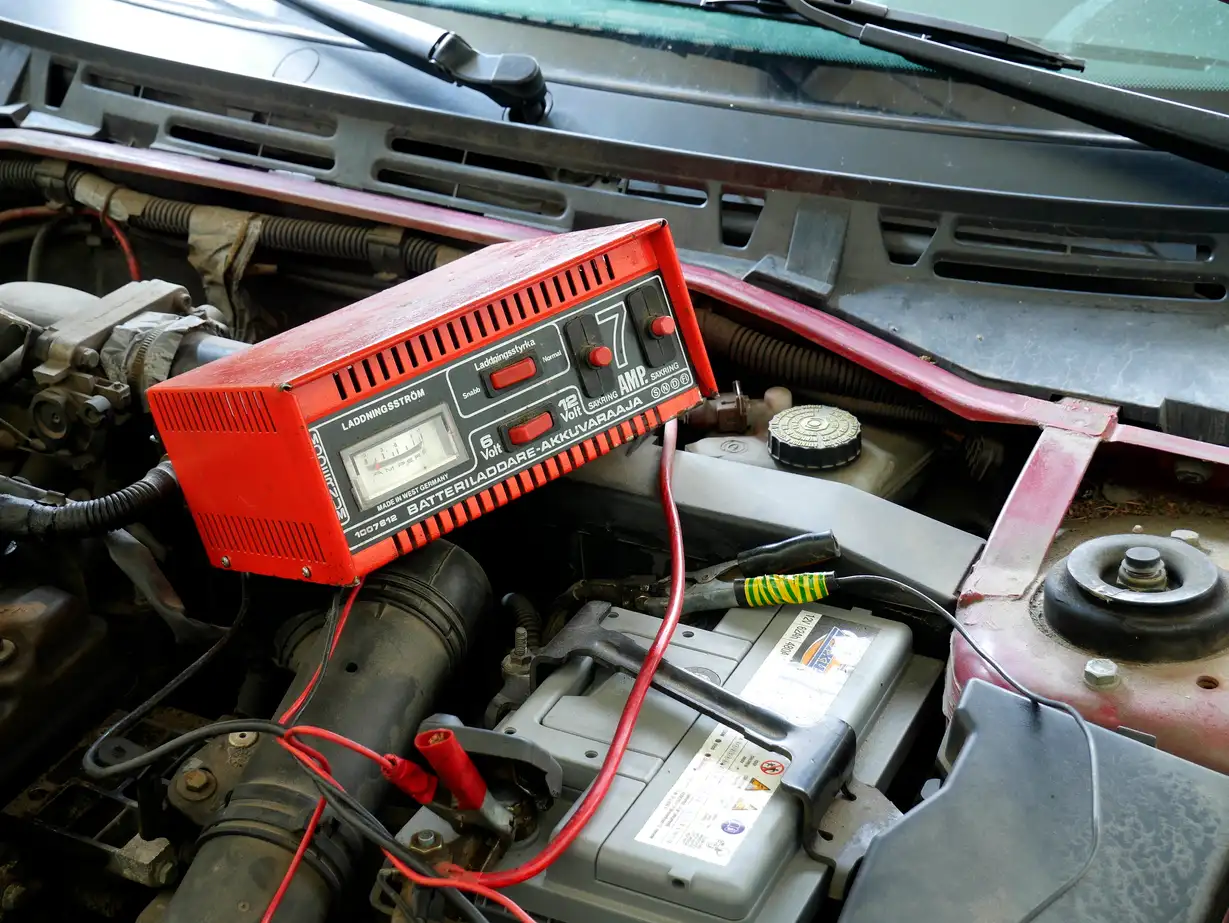 Driver beside an open-hood car holding a jump starter next to a battery charger in a driveway