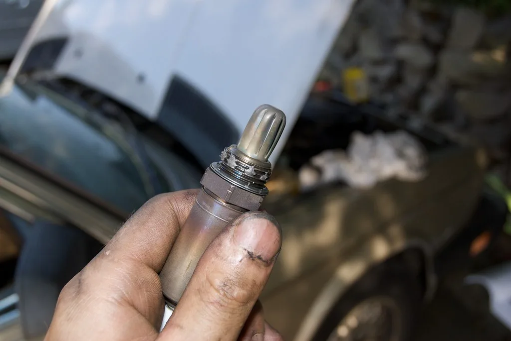 Hand holding a used oxygen sensor beside a scan tool under a lifted car