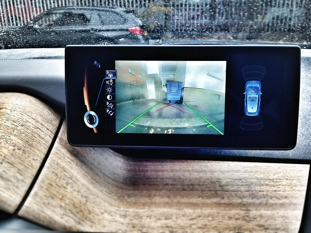 Driver backing a compact SUV into a tight city parking space with parking sensors and a 360 camera display visible