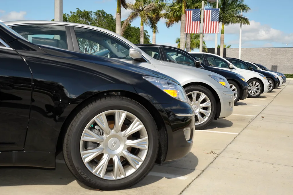Car owner compares offers beside a used sedan with a dealership lot in the background.