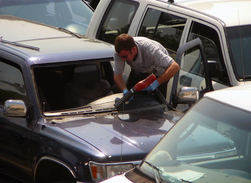 Auto glass repair technician working on a car windshield in a driveway with the customer nearby