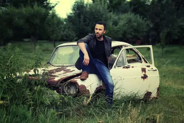 A sagging compact car sits in a salvage yard with other worn-out failed cars behind it.