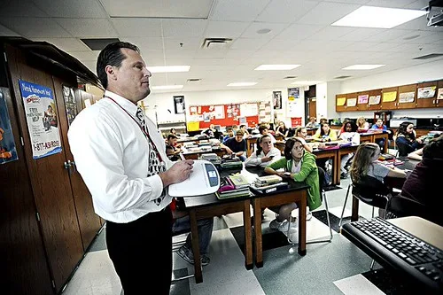 A teacher stands by tablets in a classroom while students look disengaged and the lesson plan behind them is messy.